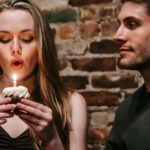 Make Friends - Young couple in elegant outfits in restaurant while blowing in candle on small cupcake and celebrating birthday near brick wall