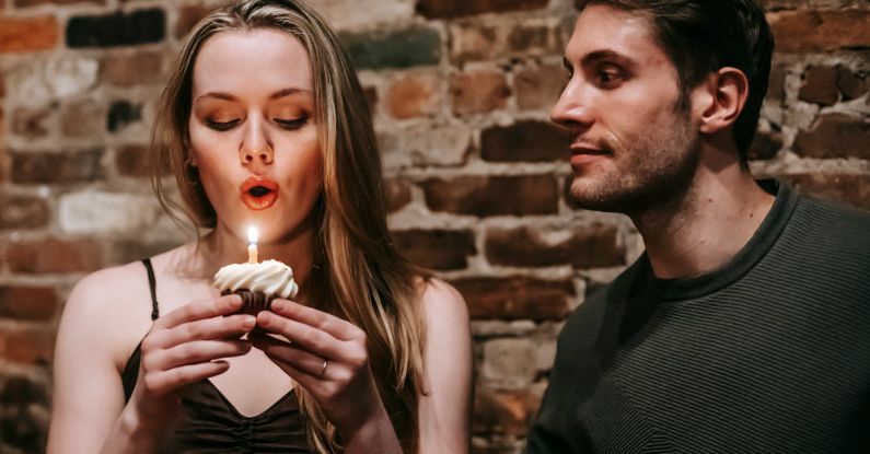 Make Friends - Young couple in elegant outfits in restaurant while blowing in candle on small cupcake and celebrating birthday near brick wall
